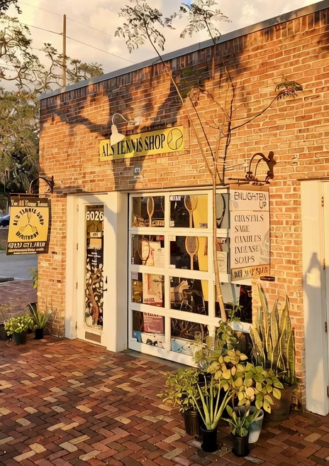 The Al's Tennis Shop storefront at golden hour. A customer walks through the door beneath a yellow 'TENNIS SHOP' sign on a warm brick building.